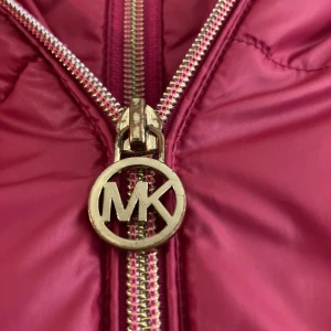 Röd pufferjacka från Michael Kors - Snygg röd pufferjacka från Michael Kors med guldig dragkedja och MK-logga. Jackan har huva, långa ärmar och quiltad design. Insidan är fodrad med mjuk fleece och har en dekorativ guldfärgad rand. Perfekt för kalla dagar och riktigt skön att bära.