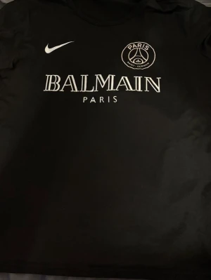 Svart PSG x Balmain t-shirt - STORLEK S. Svart t-shirt med tryck från Paris Saint-Germain och Balmain, samt Nike-logga på bröstet. Klassisk rund hals och korta ärmar. Materialet känns mjukt och lätt, perfekt för dig som gillar streetwear och sportiga vibes.