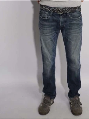 Replay Jeans blåa med slitningar  - Säljer ett par klassiska Replay jeans med raka ben och snygga slitningar. Jeansen har fem fickor, normal passform och är tillverkade i slitstarkt denim. Perfekta till vardags för en avslappnad look.