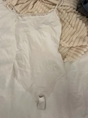 Vit spetskantad bodysuit från new yorker  - Säljer en vit bodysuit från new yorker i storlek XS. Den har tunna axelband och en snygg spetskant upptill som ger en feminin touch. Materialet är mjukt och lent, perfekt för att styla med jeans eller kjol. Bodyn har knäppning nedtill för enkel användning.