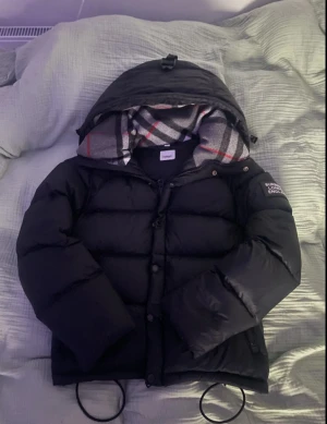 Svart dunjacka från Burberry - Säljer en svart dunjacka från Burberry med huva och klassiskt rutigt foder i grått, vitt och rött. Jackan har en patch med Burberry London England på ärmen, dragkedja och knappar framtill samt justerbara snören vid nederkant. Perfekt för kalla dagar. Hör av er vid minsta fundering. Kan tänkas gå ner i pris vid snabb affär 