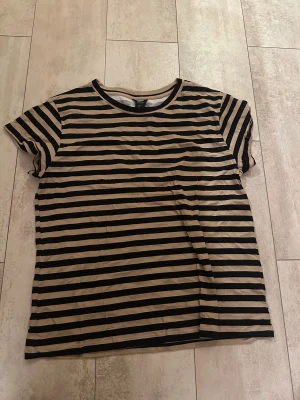 Randig beige och svart t-shirt Lindex - Säljer en beige och svart randig t-shirt från Lindex i storlek L. T-shirten har korta ärmar och rund halsringning. Materialet är mjuk bomull som känns skönt mot huden. Perfekt för dig som gillar enkel och stilren design med lite edge.