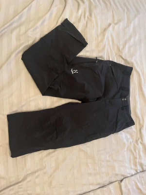 Svarta täckbyxor från Haglöfs - Svarta täckbyxor från Haglöfs med stilren design och praktiska dragkedjor vid fickorna. Byxorna har normal passform och är gjorda i slitstarkt syntetmaterial, perfekta för utomhusaktiviteter under vintern. Snyggt broderad Haglöfs-logga på benet.