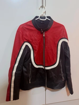 Röd och svart skinnjacka J.Lindeberg - Säljer en riktigt snygg skinnjacka från J.Lindeberg med coola röda och svarta paneler, vita detaljer och dragkedjor framtill. Jackan har en unik design med vita ränder längs ärmarna och runt bröstet, samt ståkrage med knapp. Perfekt för dig som vill sticka ut.