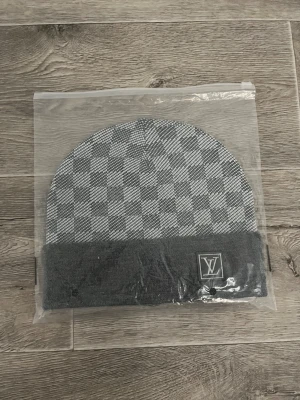Grå rutig Louis Vuitton mössa - Snygg grå mössa från Louis Vuitton med klassiskt rutigt mönster och uppvikt kant med LV-logga framtill. Mössan är stickad i 100% kashmir vilket gör den supermjuk och lyxig. Perfekt för dig som vill ha en stilren accessoar med designer-vibe.
