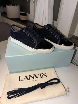 Lanvin DBB1 Lace-Up Sneakers - Snygga mörkblå sneakers från Lanvin med ovandel i mocka och vit platt sula. Skorna har snörning och en glansig svart lackad tå som ger en lyxig touch. Kommer med extra skosnören och dustbag. Perfekt för dig som gillar stilrena och exklusiva sneakers.     Skorna är köpa på FARFETCH och är endast använda ett fåtal gånger då jag råkade köpa en för stor storlek. ( har kvitto från FARFETCH). Skorna är i ett otroligt fint skick.