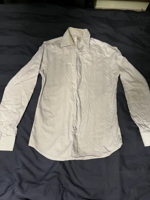 Randig skjorta från The Shirt Factory - Säljer en strykfri slim fit skjorta från The Shirt Factory i storlek 38. Skjortan är vit med tunna blå ränder, har lång ärm och klassisk krage. Knappar framtill och på manschetterna. Perfekt för dig som gillar stilrena och fräscha plagg.