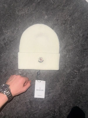 Vit ribbad Moncler mössa - Säljer en stilren vit mössa från Moncler med ribbad struktur och uppvikt kant. Mössan har Moncler-logga framtill och är tillverkad i mjukt material som håller dig varm. Perfekt accessoar för kyliga dagar och snygg till streetwear.