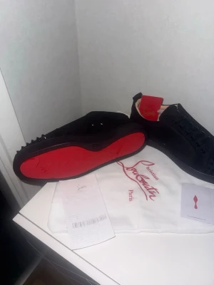 Christian Louboutin svarta sneakers strl 44 - Säljer ett par svarta sneakers från Christian Louboutin med ikonisk röd sula och svarta nitar på tån. Skorna har snörning och är tillverkade i mocka med läderfoder. Grymt snygga detaljer och lyxig vibe, perfekt för dig som gillar statement pieces.