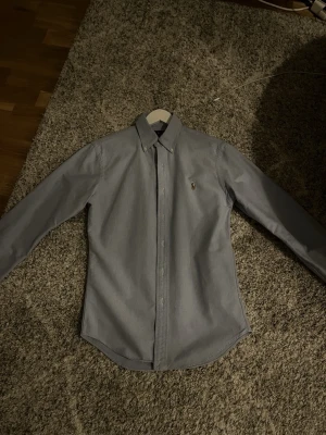 Grå slim fit skjorta från Ralph Lauren - Snygg grå skjorta från Ralph Lauren i slim fit-modell. Skjortan har klassisk krage med knappar, lång ärm och det ikoniska broderade logot på bröstet. Tillverkad i mjuk bomull som känns skön mot huden. Perfekt för dig som gillar stilrena och tidlösa plagg.