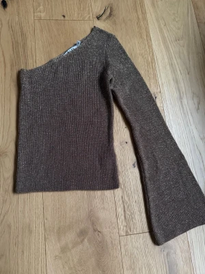 Glittrig brun oneshoulder topp från NA-KD - Säljer en brun glittrig topp från NA-KD i storlek XXS. Toppen har en snygg oneshoulder-design med lång ärm och är ribbad i materialet. Perfekt för dig som gillar att sticka ut med lite glitter och cool form.