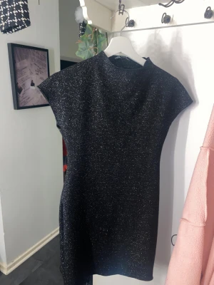 Svart glittrig kortklänning - Snygg svart kortklänning med glittriga detaljer och hög hals. Klänningen har en rak passform och är ärmlös med breda axlar. Perfekt för dig som vill sticka ut med lite extra glitter. Materialet känns mjukt och har en skimrande yta.