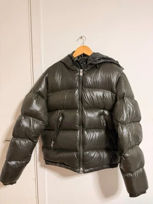 SLUTSÅLD Pufferjacka ZARA - Säljer en mörkgrön pufferjacka med glansig finish och huva. Jackan har två sidofickor med dragkedja och en kraftig dragkedja framtill. Den är quiltad och har en loose passform, perfekt för kalla dagar. Materialet är syntet och jackan är långärmad. Se näst sista bild för ”skavank”