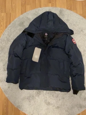 Mörkblå dunjacka från Canada Goose - Säljer en mörkblå dunjacka från Canada Goose med huva och klassisk logga på ärmen. Jackan har dragkedja och knappstängning framtill, samt ribbade muddar vid ärmslut. Perfekt för kalla dagar och riktigt snygg stil.