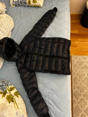  Peak Performance dunjacka - Säljer en svart dunjacka från Peak Performance i storlek 160. Jackan har huva, dragkedja framtill och två sidofickor med dragkedja. Den är quiltad och har en snygg logotyp på ärmen. Perfekt för kalla dagar och riktigt stilren.