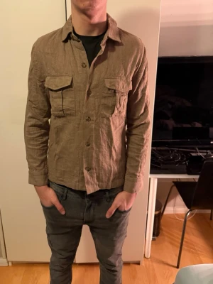 Beige overshirt i lin från Riley - Säljer en snygg beige overshirt från Riley i 100% linne. Jackan har två stora bröstfickor med lock, knäppning framtill och klassisk krage. Perfekt för lager-på-lager och ger en chill vibe till outfiten.