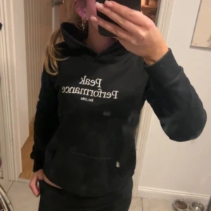 Svart hoodie från Peak Performance - Säljer en svart hoodie från Peak Performance med vit text på bröstet. Den har en klassisk design med en stor ficka framtill och en bekväm huva. Perfekt för en avslappnad stil.