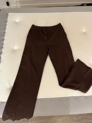 Bruna wide kostymbyxor från Monki - Snygga bruna kostymbyxor från Monki i storlek 34. Byxorna har ett wide snitt med raka, vida ben och medelhög midja. De är gjorda i mjukt bomullsmaterial och har bälteshällor, knapp och dragkedja framtill samt fickor både fram och bak.