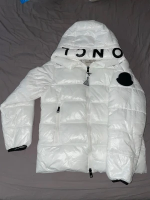 Vit pufferjacka från Moncler - Säljer en vit pufferjacka från Moncler med huva och svart logotyptryck. Jackan har dragkedja framtill, två sidofickor med dragkedja och ett svart märke på ärmen. Materialet är glansigt och jackan är dunfylld för extra värme. A1