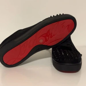 Svarta Louboutin sneakers med nitar - Säljer ett par svarta sneakers från Christian Louboutin med ikonisk röd sula och svarta nitar på tån. Skorna har snörning och är tillverkade i mocka för en lyxig känsla. Perfekt för dig som vill sticka ut med edgy detaljer och exklusiv design. Priset kan diskuteras!! En tagg saknas!!!!!