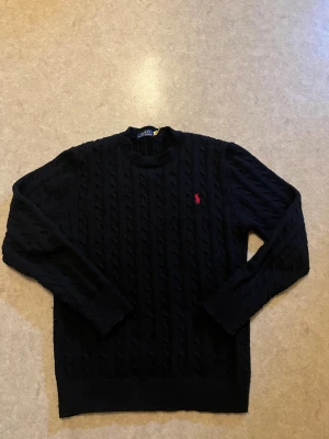 En svart stickad tröja från Polo Ralph laouren  - Snygg mörkblå stickad tröja från Polo Ralph Lauren med klassiskt kabelstickat mönster och röd broderad logga på bröstet. Rund halsringning och långa ärmar med ribbade muddar. Perfekt för dig som gillar stilrena och tidlösa plagg.