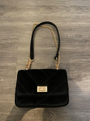 Svart quiltad axelväska med kedja - Snygg svart axelväska i quiltad design med guldfärgad kedja och vridlås framtill. Väskan är gjord i mjuk sammet och har en stilren, rektangulär form. Perfekt för dig som vill ha en trendig och elegant accessoar med lyxig känsla.