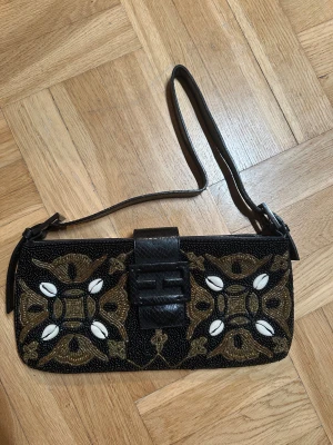 Vintage handmade  svart axelväska med pärlor - Unik svart axelväska  med detaljerad broderad design i guld och vita pärlor. Väskan har ett snyggt  i svart skinn och ett mönster som ger riktigt lyxiga vibes. Perfekt storlek för det viktigaste och en axelrem i svart skinn.