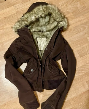 Brun bomberjacka med fuskpäls från Abercrombie & Fitch - Säljer en brun bomberjacka från Abercrombie & Fitch med mysig fuskpäls i luvan och insidan. Jackan har ribbade muddar, två fickor med knappar och dragkedja framtill. Perfekt för kalla dagar och har en cool, croppad passform.