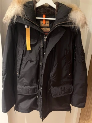 Parajumpers svart parkajacka med päls - Svart parkajacka från Parajumpers med fluffig päls på huvan, flera fickor och coola patchdetaljer på ärmen. Jackan har orange detaljer, dragkedja och knappar framtill. Perfekt för kalla dagar och har en slim fit-passning.