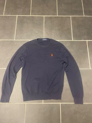 Mörkblå tröja från Polo Ralph Lauren - Mörkblå tröja från Polo Ralph Lauren i mjuk pima bomull. Klassisk rund hals och långärmad design med den ikoniska röda broderade loggan på bröstet. 