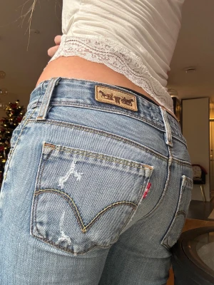 Levis 527 ljusblå bootcut jeans - Säljer mina favorit jeans från Levis 527 i ljusblå tvätt med klassiska detaljer och slitningar, då det inte passar mig längre. Modellen har låg midja och bootcut-ben, vilket ger en snygg siluett. Jeansen är gjorda i mjukt bomullsmaterial och har Levis ikoniska patch och röda etikett på bakfickan.