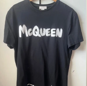 Svart Alexander McQueen t-shirt - Svart t-shirt från Alexander McQueen med vit, handmålad logotyp på bröstet. Klassisk rund hals och korta ärmar. Tillverkad i mjuk bomull och designad i Italien. Perfekt för dig som vill ha en cool och stilren look med designer-vibe.