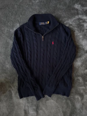Mörkblå stickad tröja Polo Ralph Lauren - Säljer en mörkblå stickad tröja från Polo Ralph Lauren i storlek XS. Tröjan har kabelstickat mönster, halv dragkedja vid kragen och en röd broderad logga på bröstet. Perfekt för kyliga dagar och har ribbade muddar vid ärmslut och nederkant.