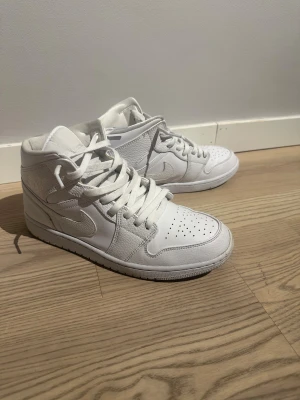 Nike Jordan 1 Mid helvita sneakers - Säljer ett par fräscha Nike Jordan 1 Mid sneakers i helvitt skinn. Klassisk siluett med perforerad tå, rund tå och snörning. Snygga och stilrena, passar perfekt till streetwear och har en clean vibe. Skorna har en platt sula och är i mid-cut modell.
