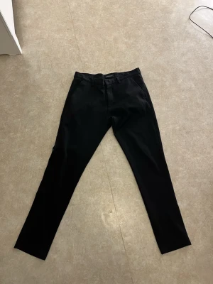 Svarta chinos från Tailored Originals W30 L30 - Svarta chinos från Tailored Originals med klassisk rak passform och diskreta fickor både fram och bak. Byxorna har bälteshällor och är gjorda i mjuk bomull som känns skön mot huden. Perfekt för dig som gillar stilrena och enkla plagg.