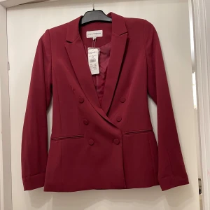 Vinröd kavaj från KappAhl, strl 34 - Snygg vinröd kavaj från KappAhl med dubbelknäppning och klassisk krage. Kavajen har långa ärmar, två fickor fram och är fodrad med matchande tyg. 