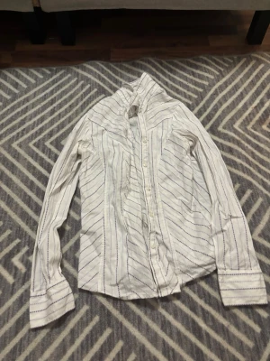 Vit randig skjorta med knappar - Snygg vit skjorta med tunna blå ränder och diskreta prickar. Skjortan har klassisk krage, långa ärmar och knäppning framtill. 