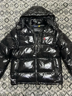 Svart pufferjacka från Polo Ralph Lauren - Snygg svart pufferjacka från Polo Ralph Lauren med glansig finish och huva. Jackan har dragkedja framtill, flera fickor med dragkedja och röd broderad logga på bröstet. Perfekt för kalla dagar och har en riktigt modern vibe.