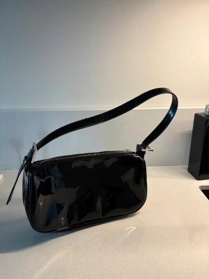 Svart axelväska från Gina Tricot - Snygg svart axelväska från Gina Tricot i glansigt syntetmaterial. Väskan har en justerbar rem med silverfärgat spänne och dragkedja upptill. Köpt för något år sedan men ingen skada på varan då den knappt används☺️