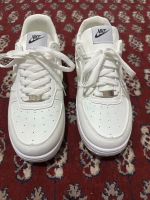 Nike Air Force 1 vita sneakers - Säljer ett par klassiska Nike Air Force 1 i helvitt skinn med vit sula och snörning. Skorna har perforerad tå, silverfärgad detalj vid snörningen och ikonisk Nike swoosh på sidan. Perfekta för dig som gillar en clean och tidlös look.