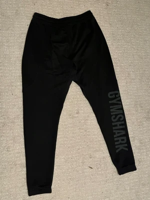 Svarta Gymshark mjukisbyxor med tryck - Svarta mjukisbyxor från Gymshark med stort tryck längs ena benet och logga vid fickan. Byxorna har snörning i midjan, ribbade muddar och är gjorda i mjukt syntetmaterial. Perfekta för gymmet eller chill hemma.