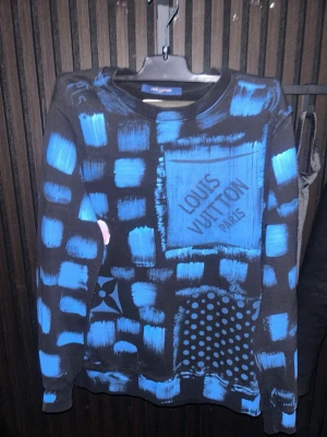 Louis Vuitton blå sweatshirt - Cool svart sweatshirt från Louis Vuitton med blått och rosa abstrakt mönster och tryck. Tröjan har lång ärm, rund hals och unikt rutigt och prickigt print med logga på både fram- och baksida. Perfekt statement-plagg för dig som gillar streetwear och designer.Finns även en Scan kod som skickar dig till Louis Vuittons hemsida.