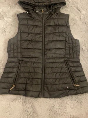 Svart dunväst från Zara - Svart dunväst från Zara med huva och två fickor med dragkedja. Västen har ett quiltat mönster och stängs med dragkedja framtill. Perfekt att slänga över en hoodie när det är kyligt ute. Snygg och enkel design som funkar till många stilar. Kan eventuellt tänka mig gå ner i pris