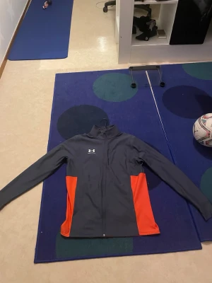 Under aroumer dress - Bilderna visar träningskläder från Under Armour, inklusive en svart och orange zip-jacka samt blåa träningsbyxor. Aldrig använd helt ny 