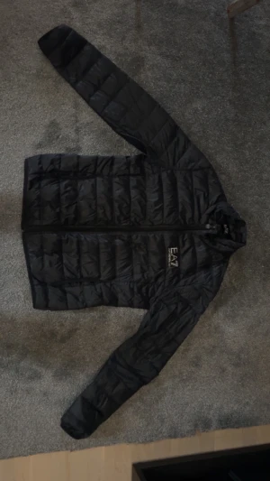 Svart pufferjacka från EA7 Emporio Armani - Svart pufferjacka från EA7 Emporio Armani med stilren design och diskret logga på bröstet. Jackan har lång ärm, dragkedja framtill och quiltad look. Materialet är polyester, vilket gör den lätt och bekväm. Perfekt för dig som vill ha en snygg och funktionell jacka.