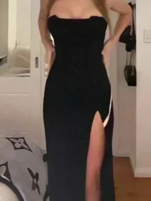 Svart glittrig långklänning med slits OH POLLY - Säljer en svart axelbandslös långklänning med glittriga detaljer. Klänningen har en snygg slits på sidan och en figurnära passform som framhäver siluetten. Materialet är mjukt och har en glittrig yta som ger extra wow-effekt. Perfekt för dig som vill sticka ut. Uk 8 storlek S