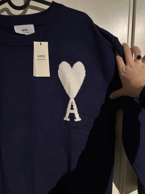 Marinblå stickad tröja från Ami - Snygg marinblå stickad tröja från Ami med vit hjärt- och A-logga på bröstet. Tröjan har rund hals, ribbade muddar och lång ärm. Perfekt för dig som gillar stilrena detaljer och vill ha något som sticker ut lite extra.