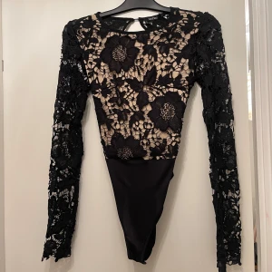 Svart spetsbody från Boohoo - Säljer en svart body från Boohoo med långärmade spetsärmar och blommigt broderat mönster. Toppen har öppen rygg med knyt och transparent mesh i beige underton. Perfekt för dig som vill ha en statement piece med snygga detaljer.