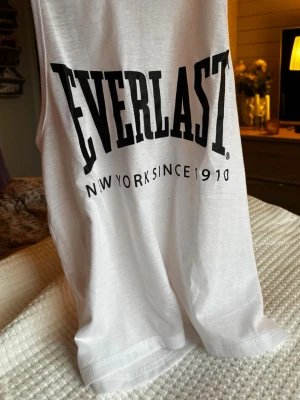 Vit linnetopp från Everlast - Säljer en vit linnetopp från Everlast med stort svart tryck på bröstet där det står 'Everlast New York Since 1910'. Toppen är ärmlös och har rund halsringning. Perfekt för träning eller chill dagar. Materialet är mjuk bomull som känns skönt mot huden.