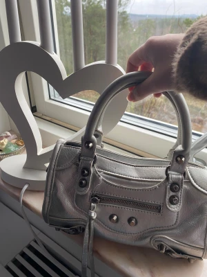 Silver handväska med dragkedja - Cool handväska i metallic silver med två rejäla handtag och snygga metalldetaljer. Väskan har en praktisk dragkedja framtill och dekorativa nitar. Tillverkad i skinnimitation som ger en lyxig känsla. Perfekt storlek för det viktigaste och riktigt trendig look.